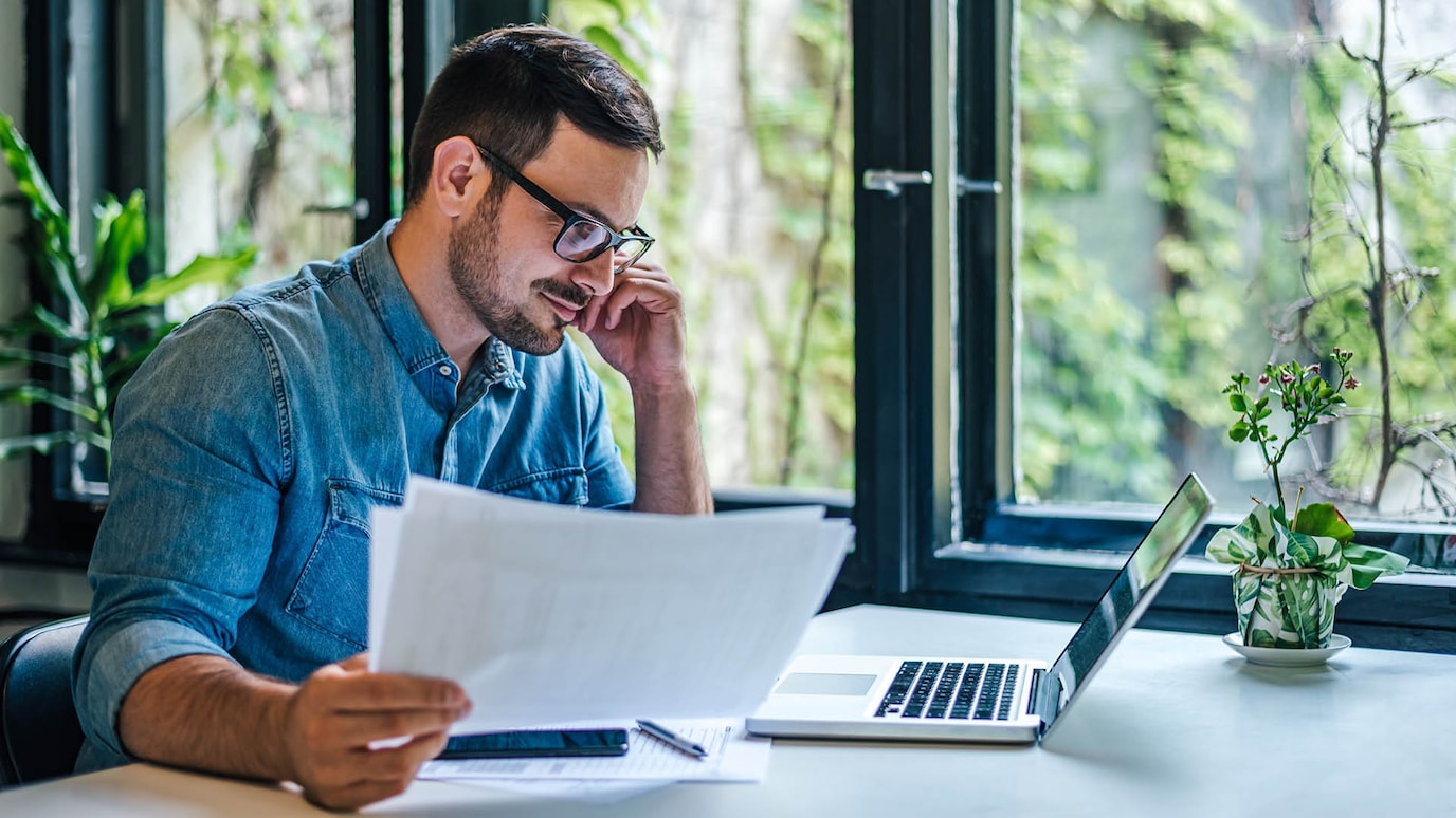 A person holding documents in their hand and looking at their laptop screen.