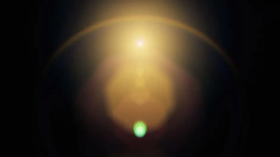 Abstract image of a bright golden light source with a halo effect against a dark background. A small green orb is visible at the bottom center, creating a contrast with the warm tones above.